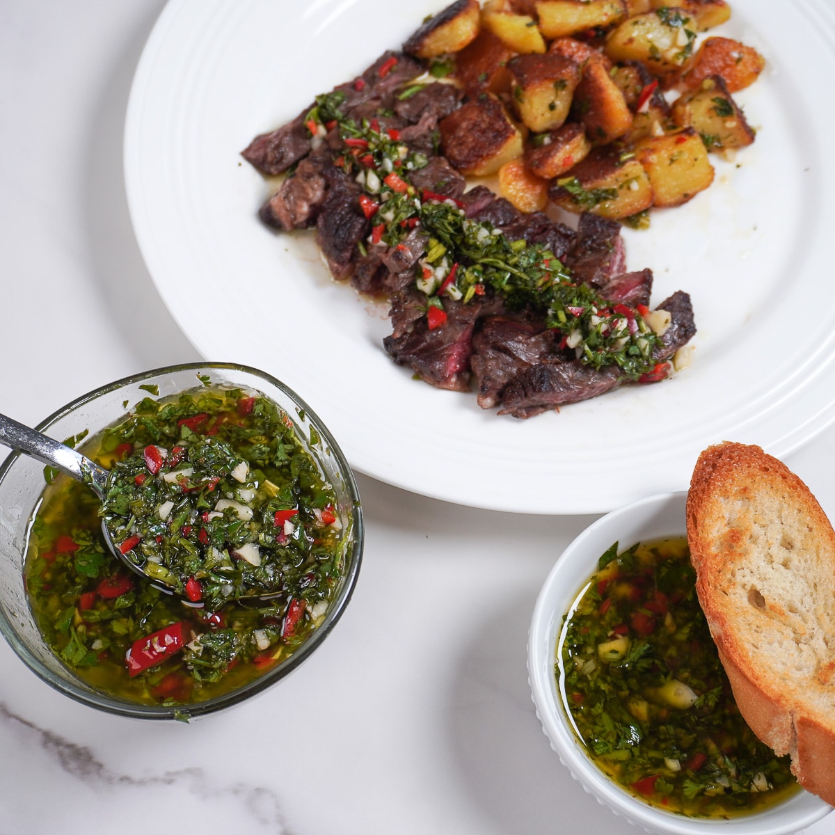 A plate filled with steak and potatoes with chimichurri sauce drizzled on top. A bowl of chimichurri sauce sits to the side of the plate.