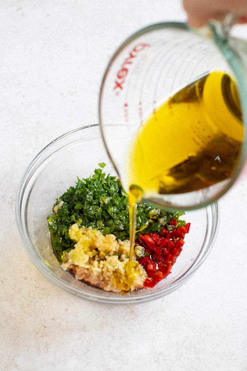 Olive oil being poured into a bowl with fresh chopped parsley, minced garlic, and red pepper.