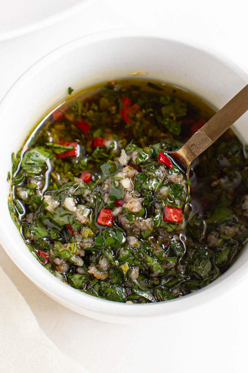 A small white bowl filled with chimichurri sauce. A spoon is scooping out some of the sauce from the bowl.