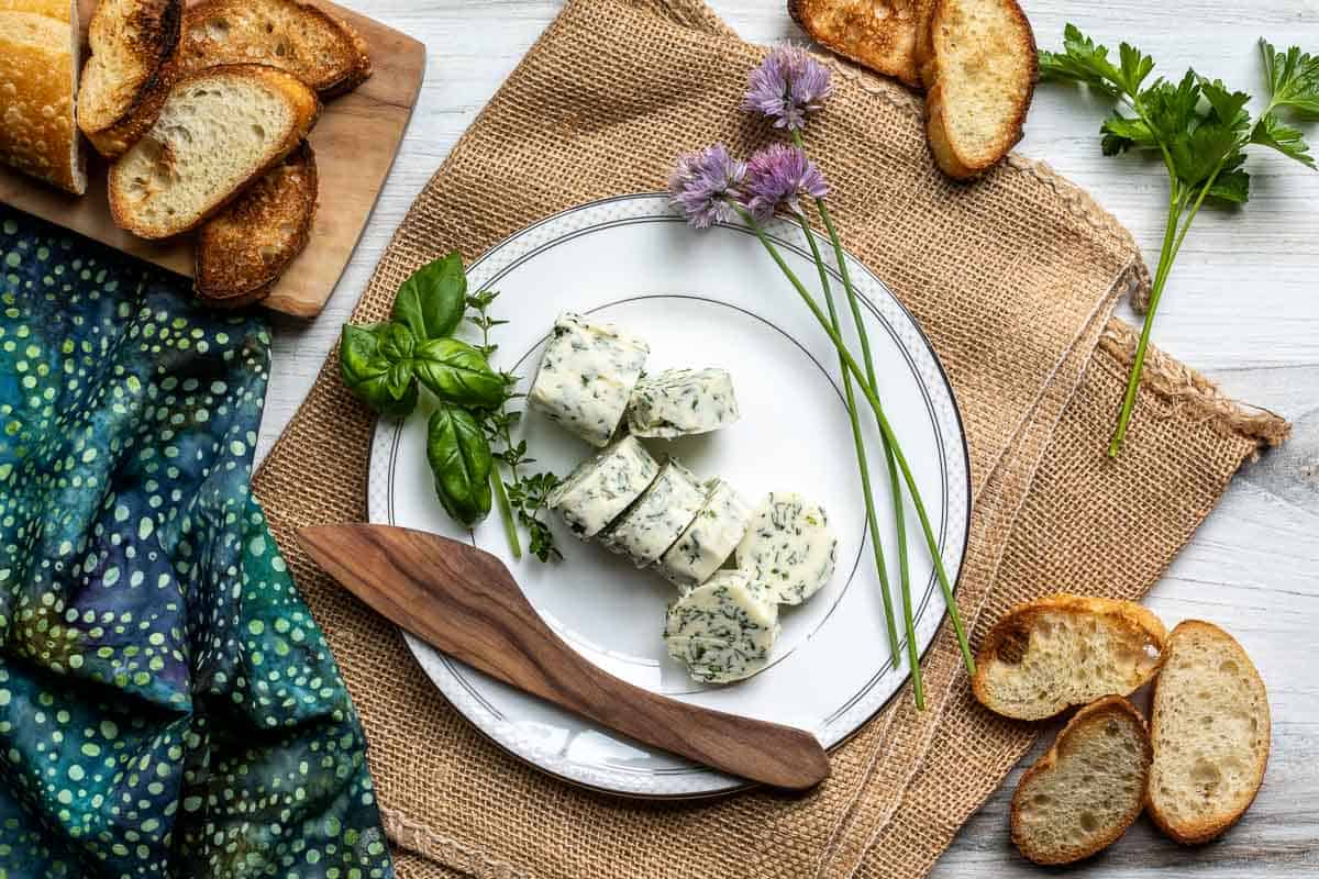 Sliced herb butter on a white plate with fresh herbs, wooden spreader, and toasted bread slices.