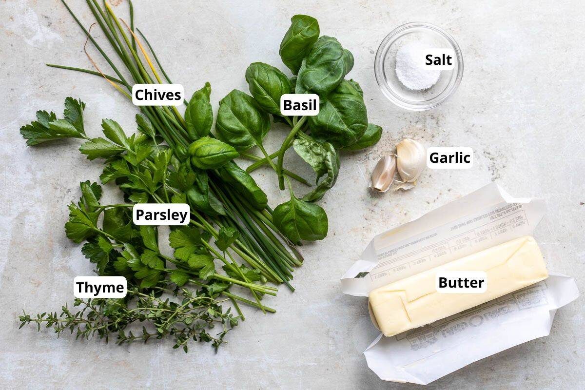 Labeled ingredients for herb compound butter: chives, basil, parsley, thyme, salt, garlic cloves, and a stick of butter on a light surface.
