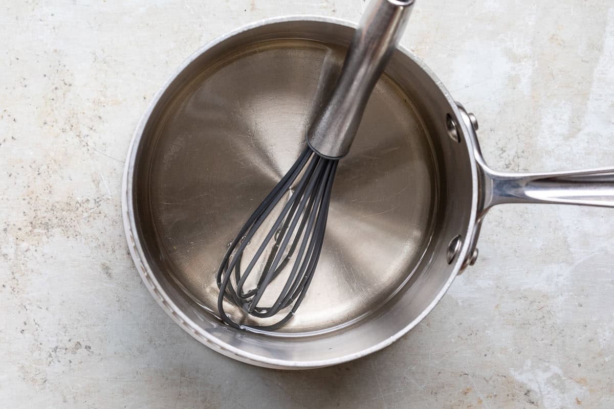 Sugar and water are being heated in a saucepan with a whisk nearby.