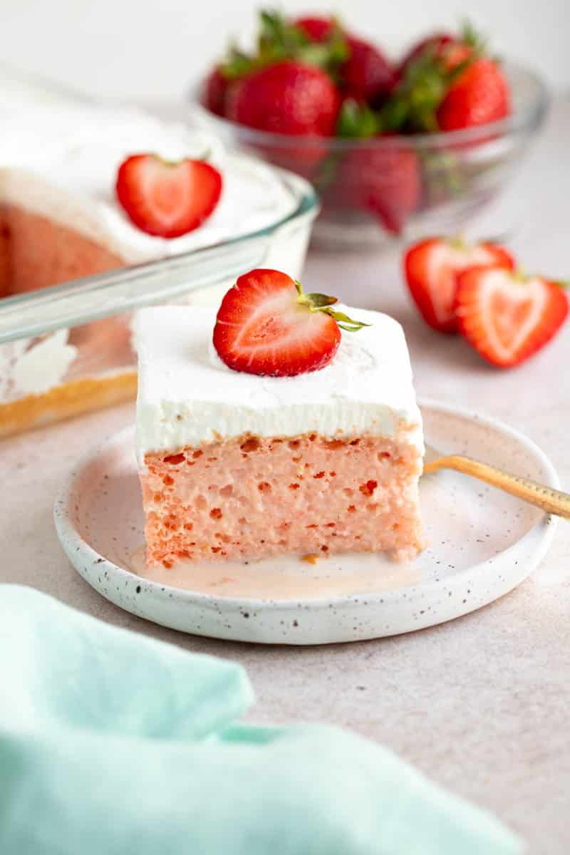 A slice of strawberry tres leches cake on a white plate, with sliced strawberries and a bowl of strawberries on a countertop.