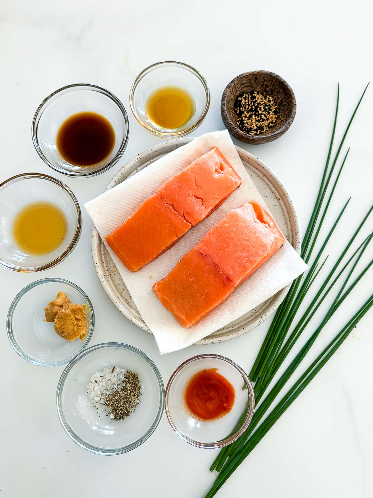 Ingredients for broiled salmon laid out: salmon filets, sesame seeds, sesame oil, soy sauce, olive oil, miso paste, salt, pepper, and sriracha, next to chives.