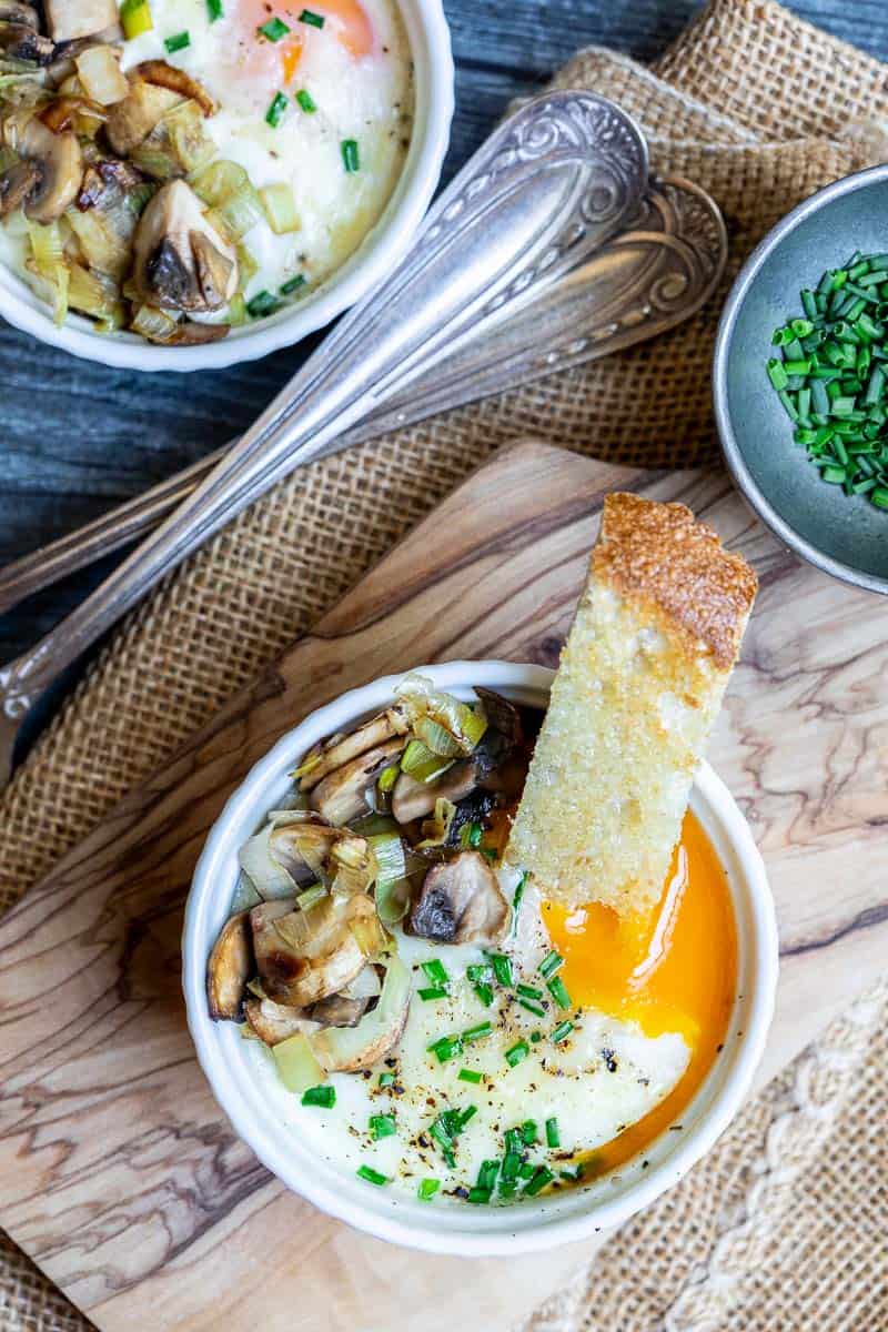 Oeufs Cocotte topped with mushrooms and chives in a ramekin beside toasted bread.